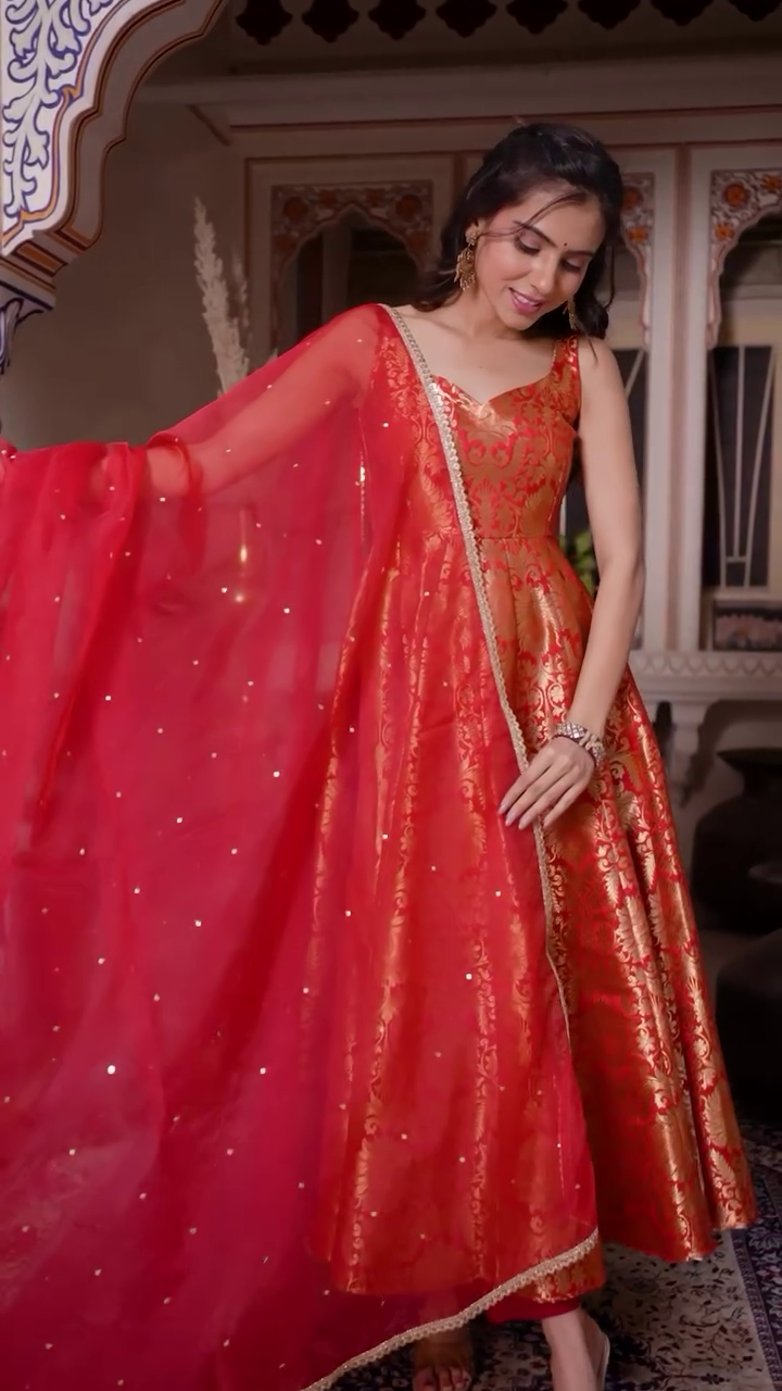 Gracefully Posing in a Stunning Red Anarkali Dress with Intricate Golden Embroidery, Complemented by a Sheer Red Dupatta Adorned with Sparkling Details, Radiating Elegance for a Traditional Celebration