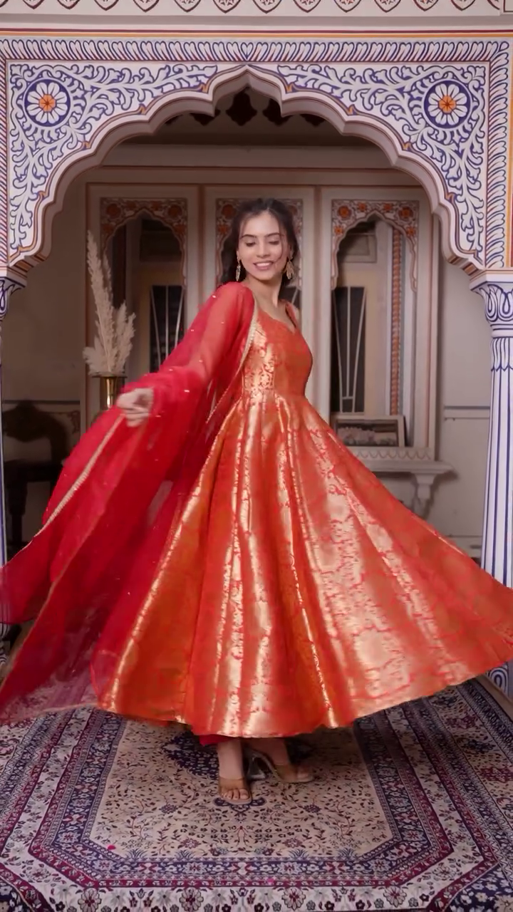 Gracefully Posing in a Stunning Red Anarkali Dress with Intricate Golden Embroidery, Complemented by a Sheer Red Dupatta Adorned with Sparkling Details, Radiating Elegance for a Traditional Celebration
